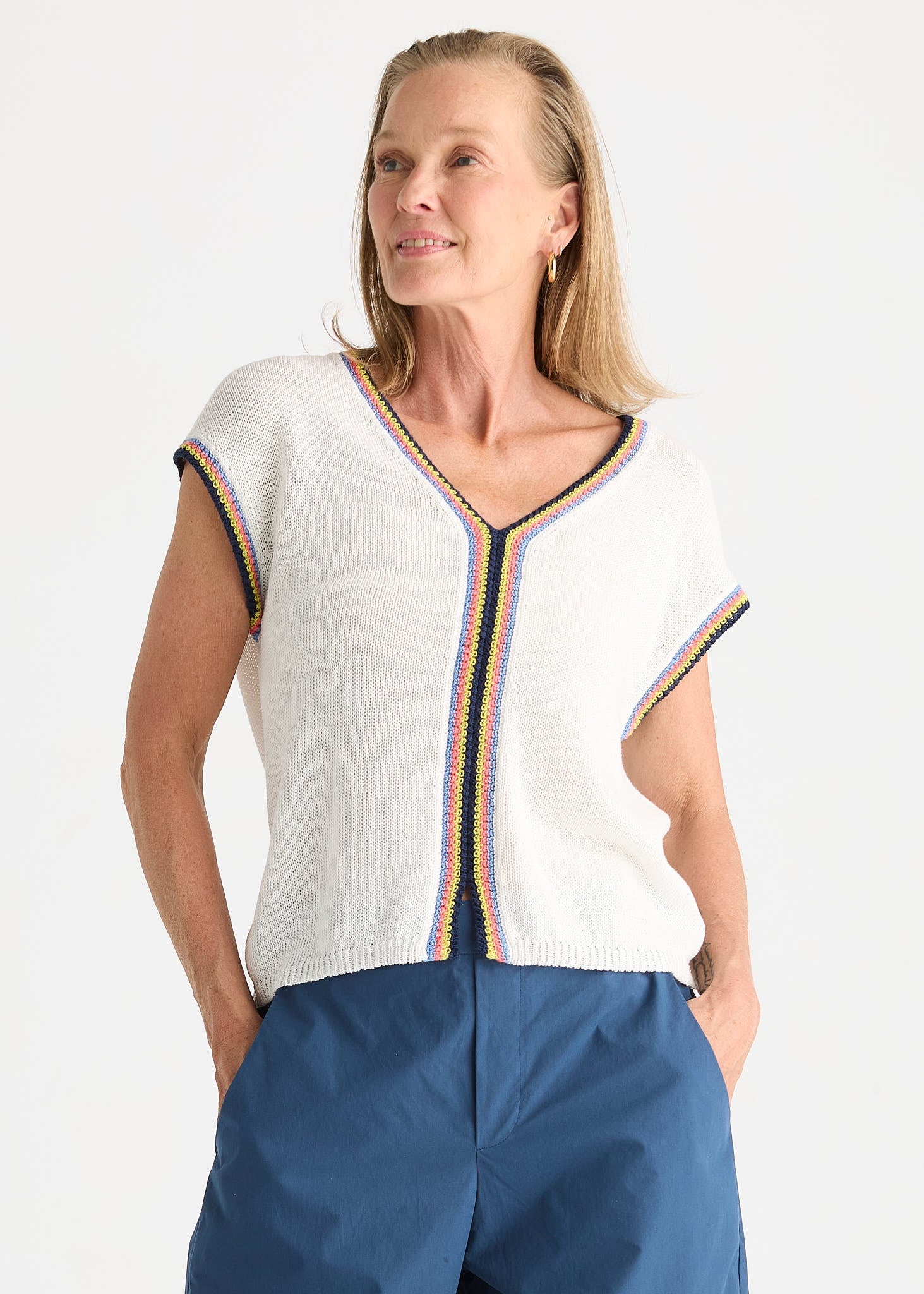 A woman wearing a white knit short-sleeve top with colorful striped trim and blue pants, photographed against a plain light background.