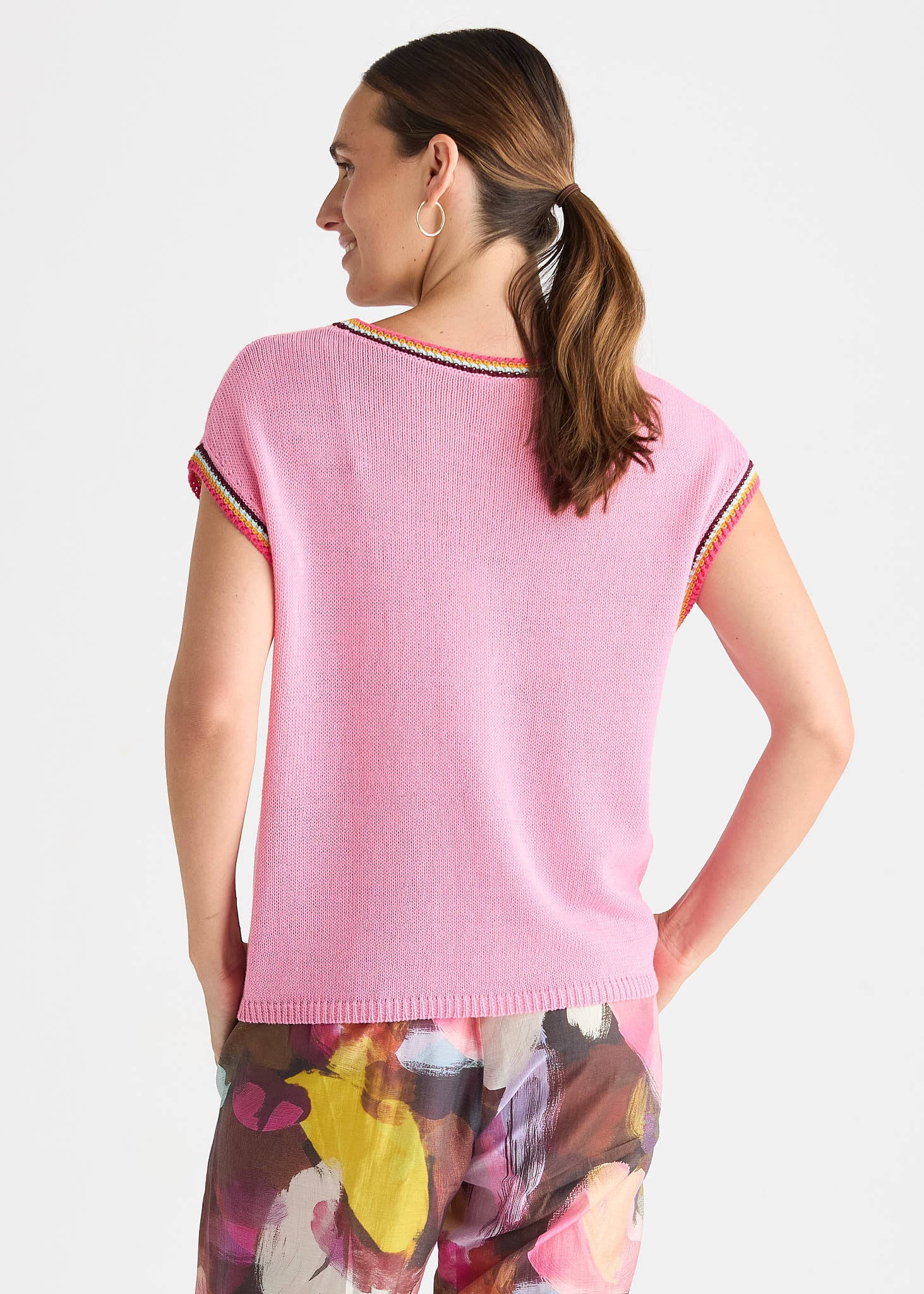 A woman shown from the back wearing a light pink short-sleeve knit top with thin multicolor striped trim along the neckline and sleeve cuffs, paired with floral print pants against a light gray studio background.