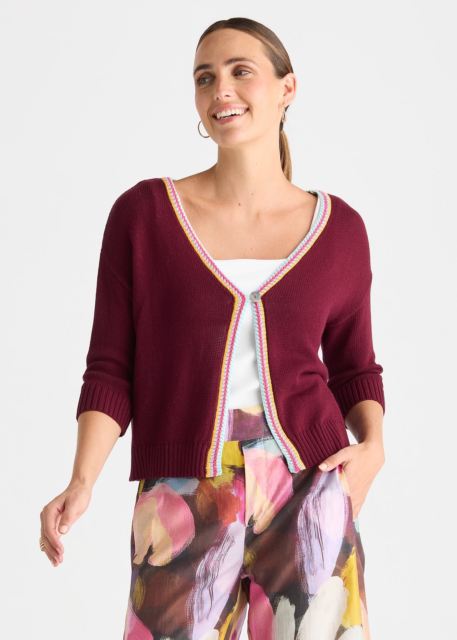 A woman wearing a burgundy knit cardigan with a colorful striped trim over a white top and floral print pants, photographed against a light gray studio background.