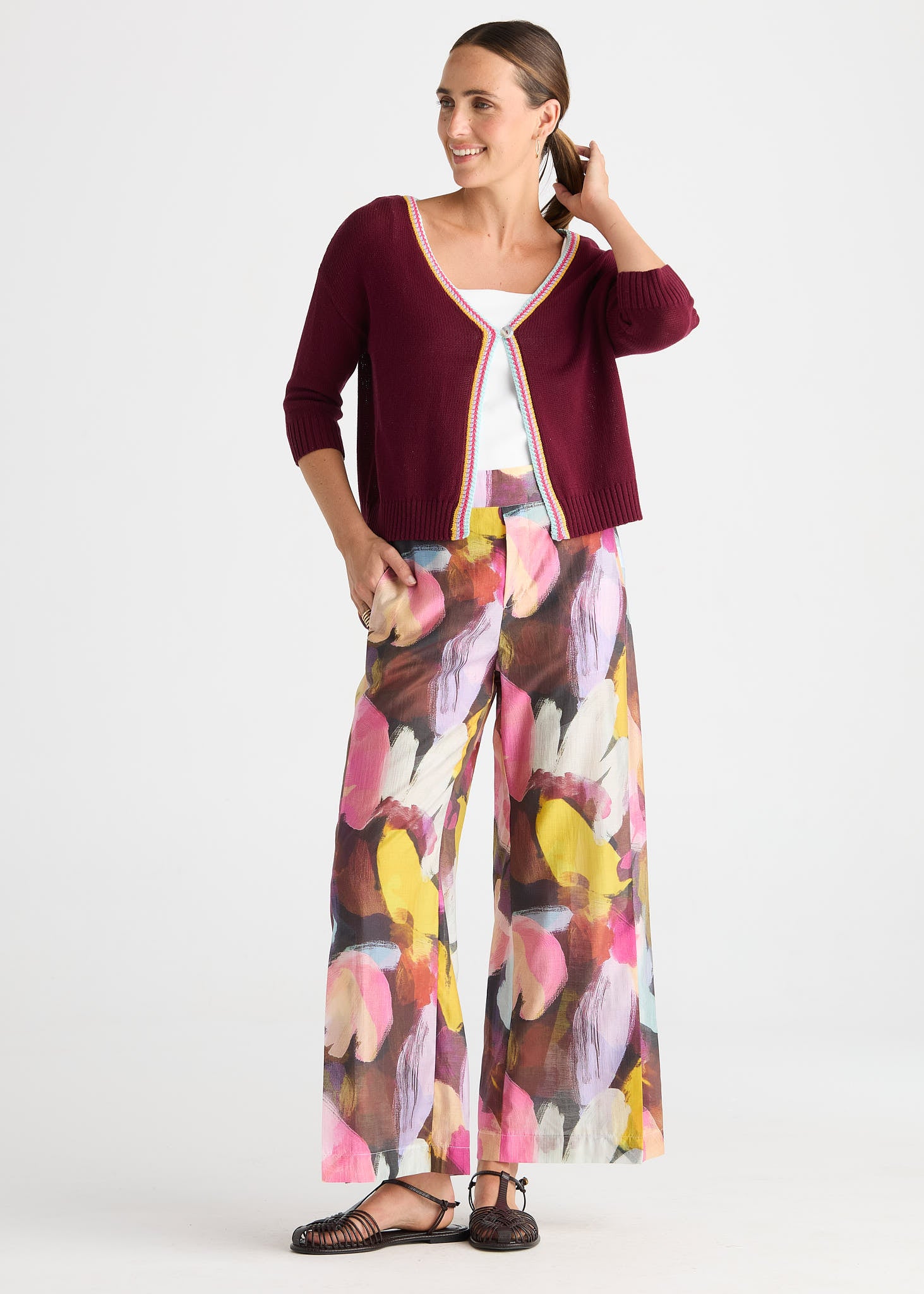 A woman modeling a three-piece outfit consisting of a burgundy cardigan with cream trim, a white camisole, and wide-leg pants with a colorful abstract floral print in pink, yellow, brown, and white.
