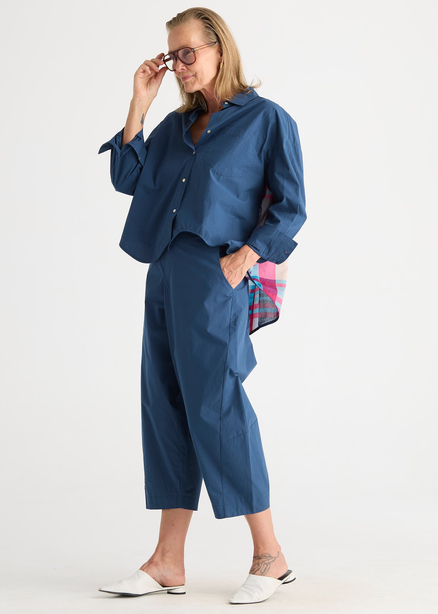 A woman modeling a matching navy blue two-piece set consisting of a cropped button-down shirt and wide-leg cropped pants, styled with white mule sandals and a pink plaid clutch bag against a plain white studio background.