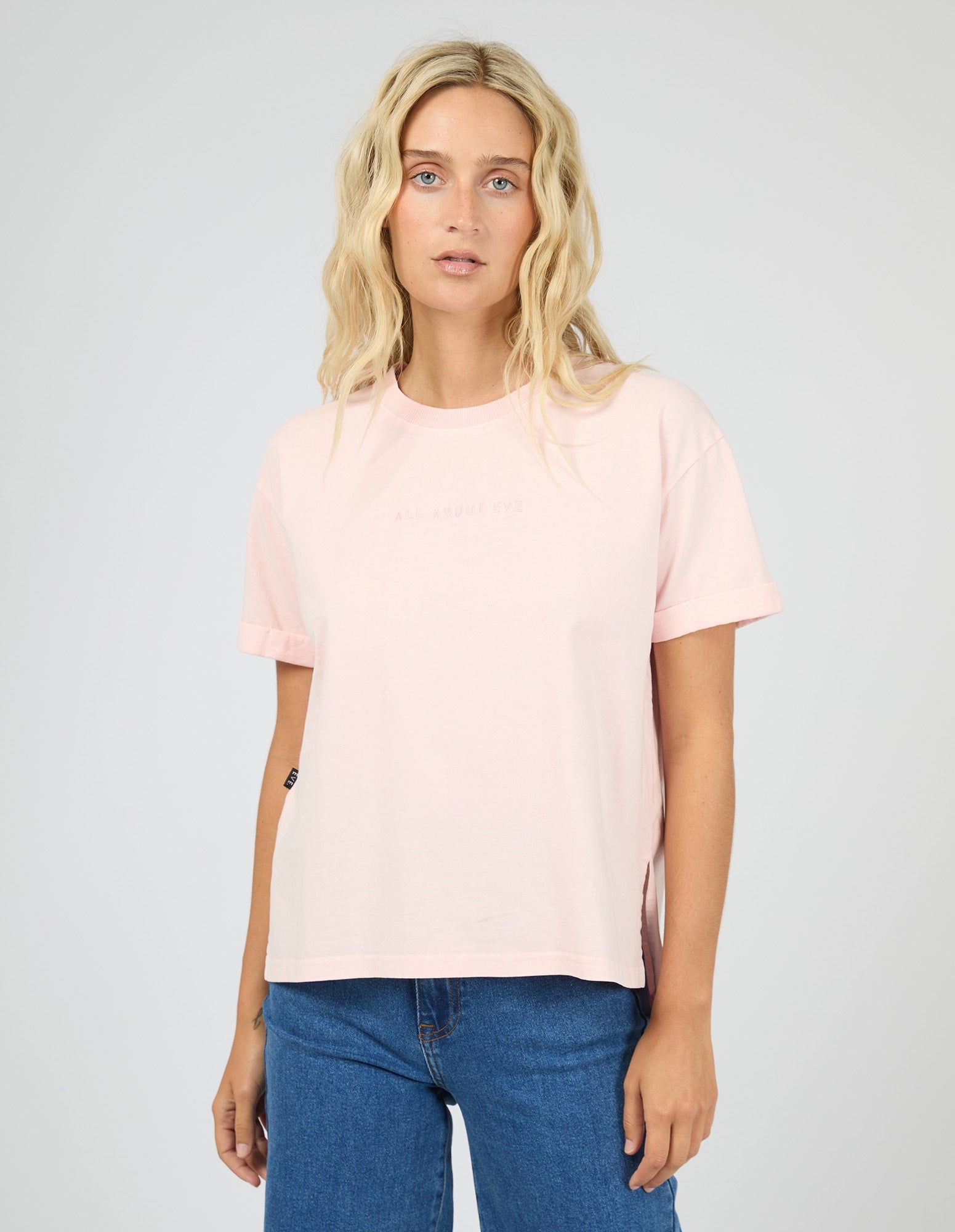 A blonde woman wearing a light pink short-sleeve crewneck t-shirt and blue jeans, standing against a light gray studio background.