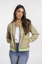 A woman modeling a green patterned bomber jacket with striped ribbed trim, worn over a white top and blue jeans, against a light gray studio background.