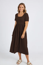 A woman modeling a short-sleeve brown midi dress with a square neckline and a gathered skirt, paired with beige sandals, against a plain light gray studio background.