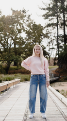 Person wearing a pink sweater and blue jeans standing on a wooden pathway with trees in the background