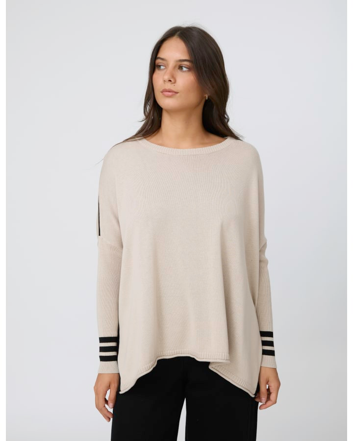 A woman wearing a light beige knit sweater with black and white striped cuffs, paired with black pants, against a light gray studio background.