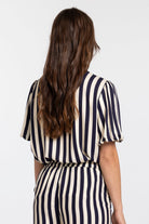 A woman viewed from behind wearing a cream and navy blue vertically striped jumpsuit with short puff sleeves and a tie waist, against a plain white background.