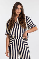 A woman modeling a short-sleeve collared jumpsuit with vertical navy and cream stripes, photographed against a clean light gray studio background.