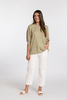 A woman modeling a loose-fitting, short-sleeved top with a green, yellow, and orange geometric pattern, paired with white cropped pants and light-colored slide sandals, against a plain light gray studio background.