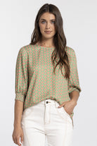 A woman modeling a green three-quarter sleeve top with a red and yellow geometric pattern, paired with white pants, against a light gray background.