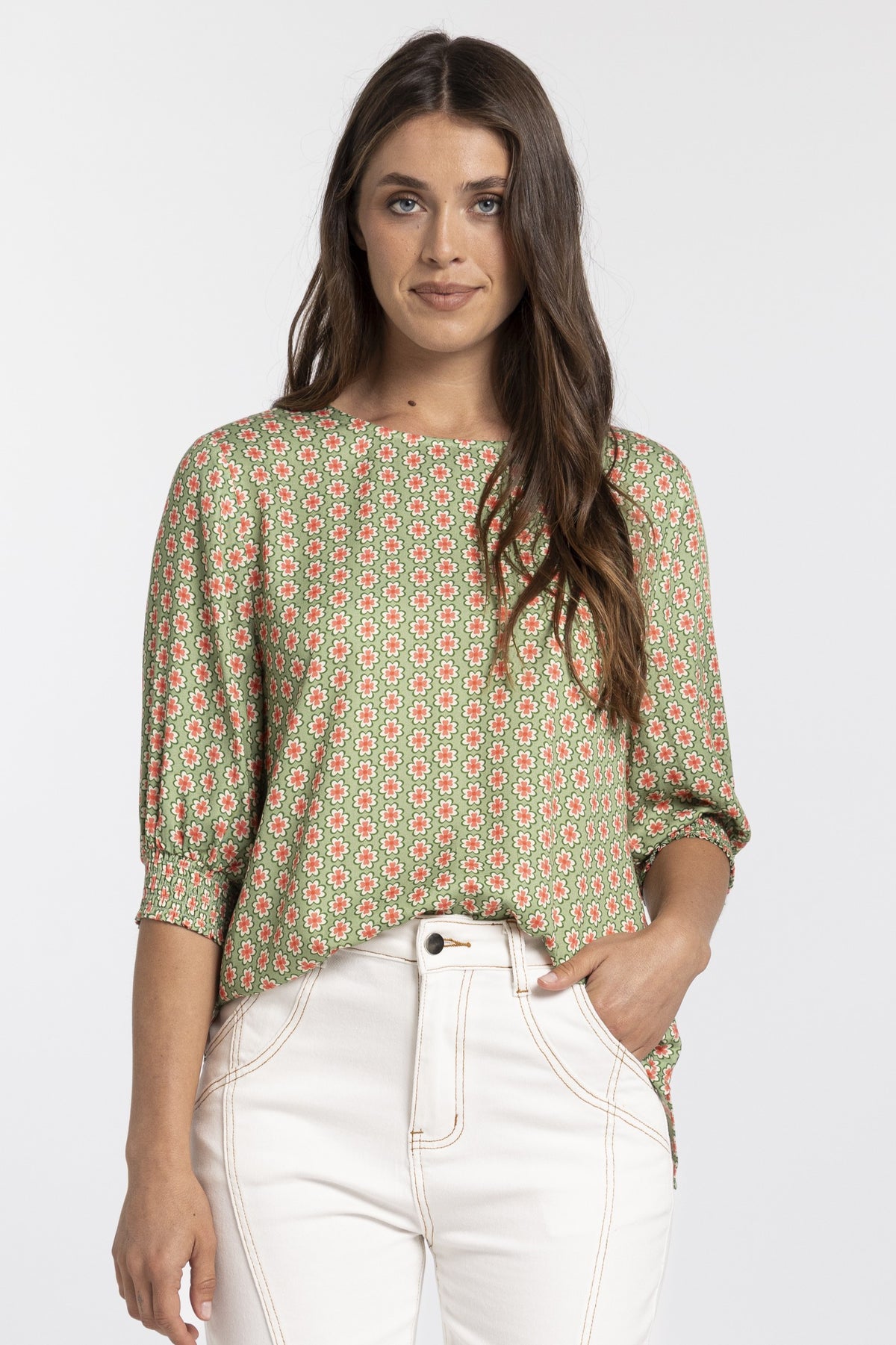 A woman modeling a green three-quarter sleeve top with a red and yellow geometric pattern, paired with white pants, against a light gray background.