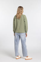 A woman is shown from behind wearing an olive green long-sleeve button-up shirt, light-wash blue jeans, and cream-colored sneakers against a plain light gray studio background.