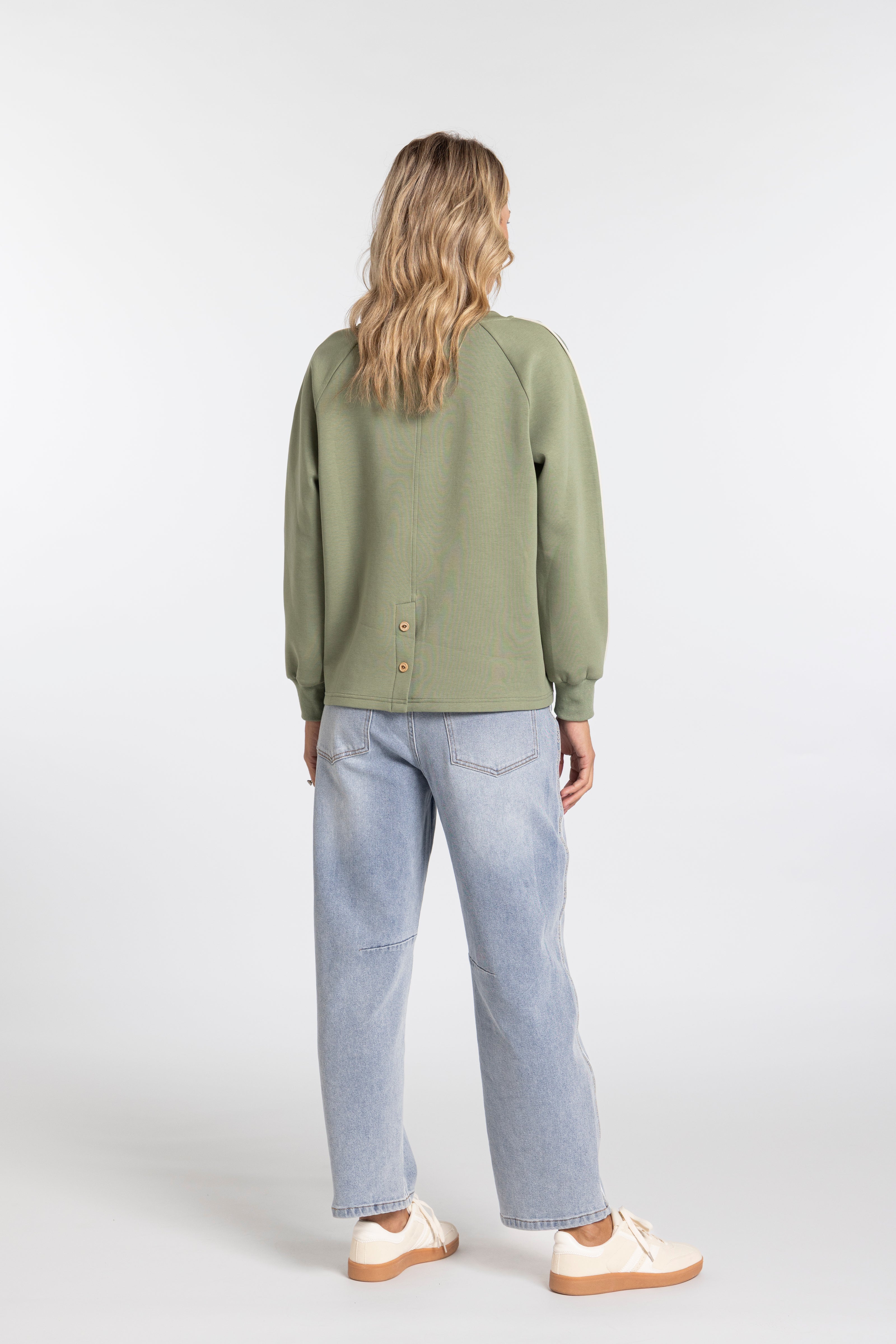 A woman is shown from behind wearing an olive green long-sleeve button-up shirt, light-wash blue jeans, and cream-colored sneakers against a plain light gray studio background.