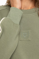 Close-up of a person wearing an olive green crewneck sweatshirt, showing the ribbed collar, sleeve cuff, and a tonal embroidered chest patch.