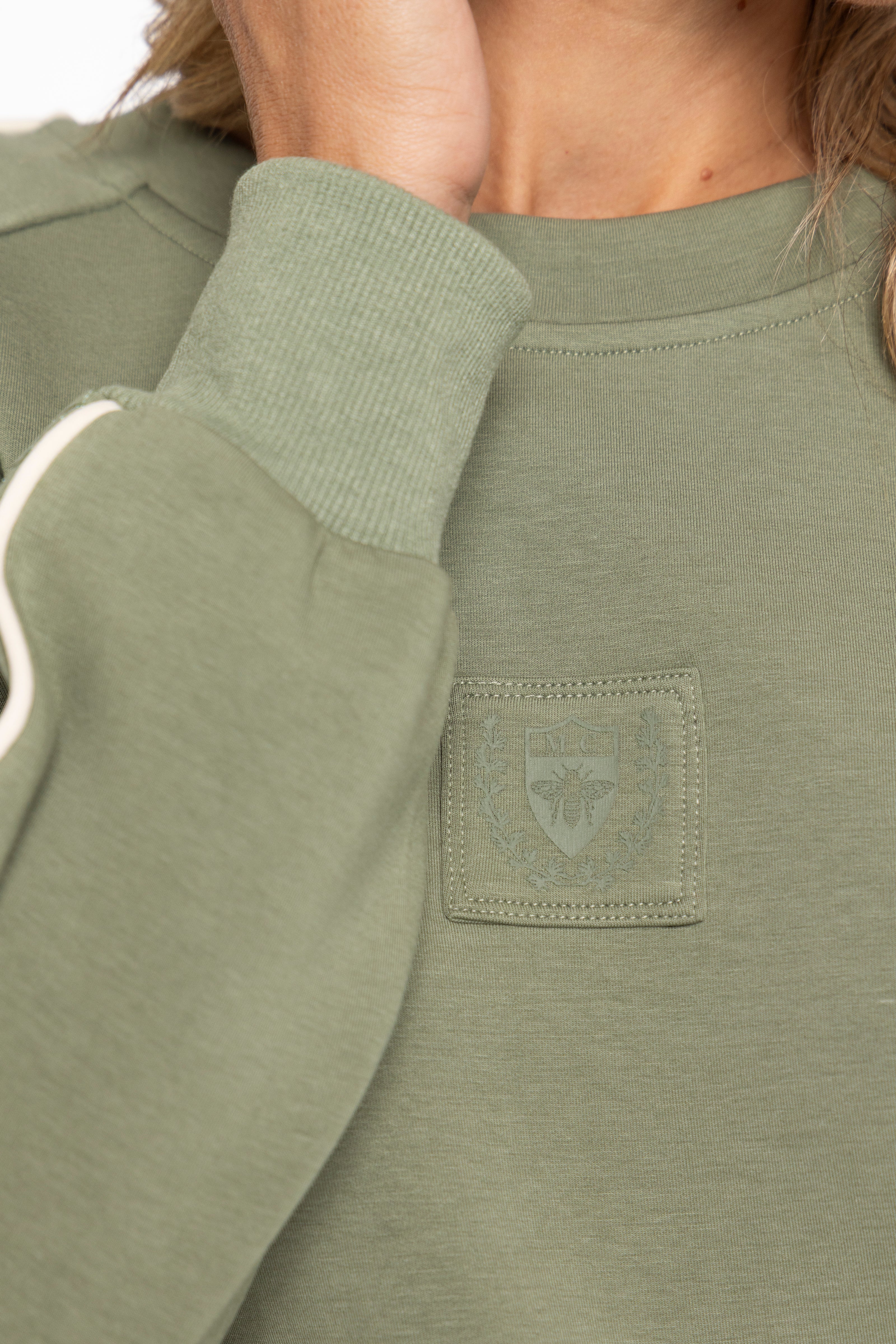Close-up of a person wearing an olive green crewneck sweatshirt, showing the ribbed collar, sleeve cuff, and a tonal embroidered chest patch.