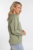 A woman wearing an olive green long-sleeve sweatshirt with white contrast piping down the sleeves, paired with light blue jeans, photographed against a plain white studio background.