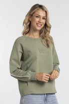 A woman wearing an olive green long-sleeve sweatshirt with white contrast stitching on the sleeves and a tonal chest pocket, paired with light blue jeans against a light gray studio background.