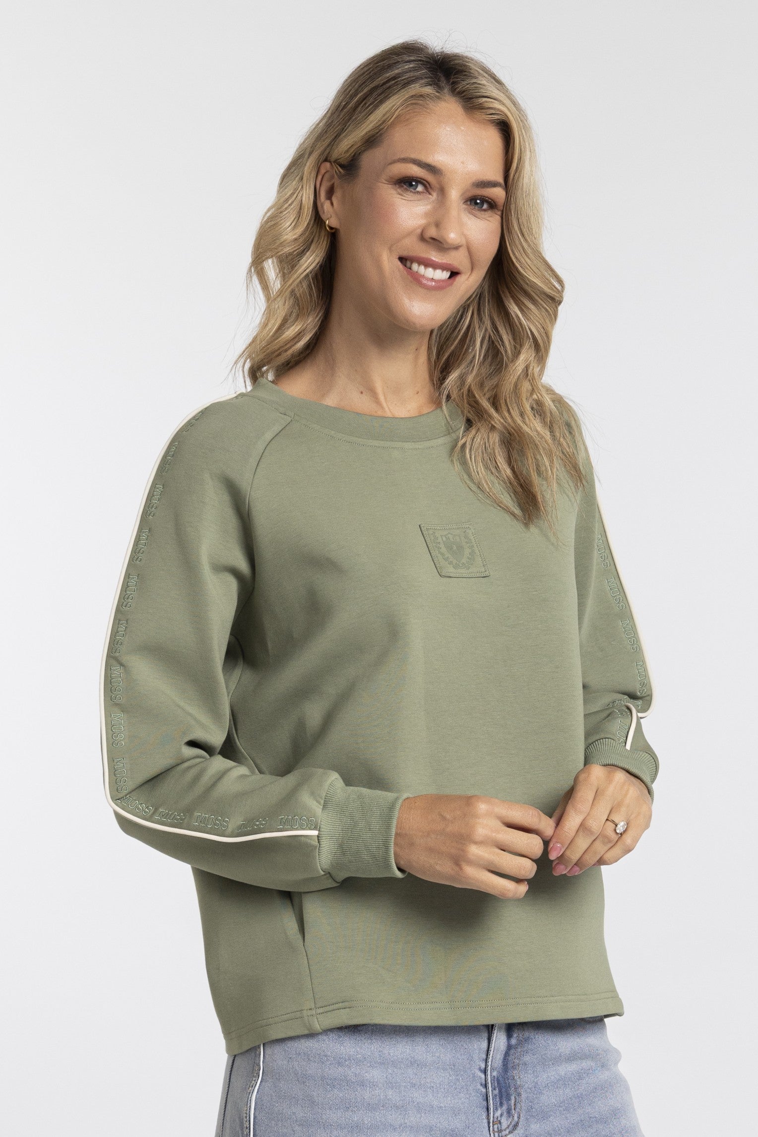 A woman wearing an olive green long-sleeve sweatshirt with white contrast stitching on the sleeves and a tonal chest pocket, paired with light blue jeans against a light gray studio background.