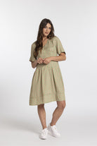 A woman modeling a short-sleeve midi dress with a dense multicolor geometric pattern on a light sage green base, paired with white sneakers, against a plain light gray studio background.