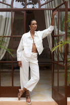 Woman in a white outfit standing in front of a glass door with plants around.