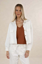 Woman wearing a white jacket, brown top, and white pants against a beige background