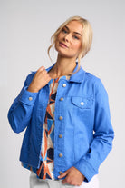 A blonde woman wearing a bright blue denim jacket over a patterned top, posing against a light gray studio background.