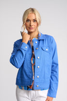 A blonde woman wearing a bright blue denim jacket over a multicolored patterned top and white pants, photographed against a light gray studio background.