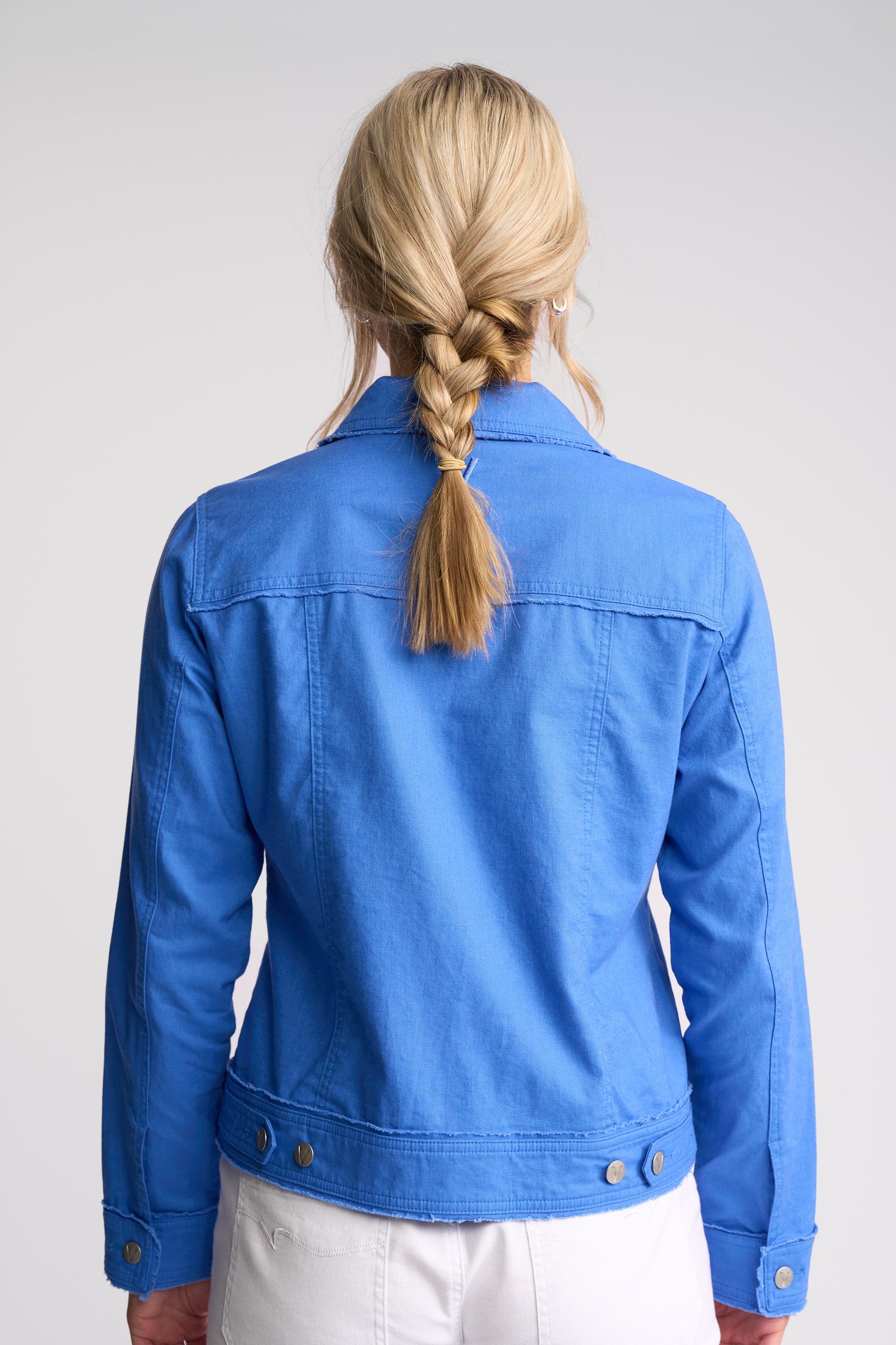 A woman viewed from behind wearing a bright blue cropped denim jacket and white pants against a light gray studio background.