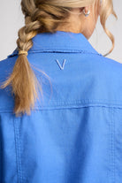 Close-up back view of a person wearing a bright blue collared shirt or jacket, with a small embroidered V-shaped logo centered below the collar. The person has blonde hair in a braid and is shown against a light neutral background.
