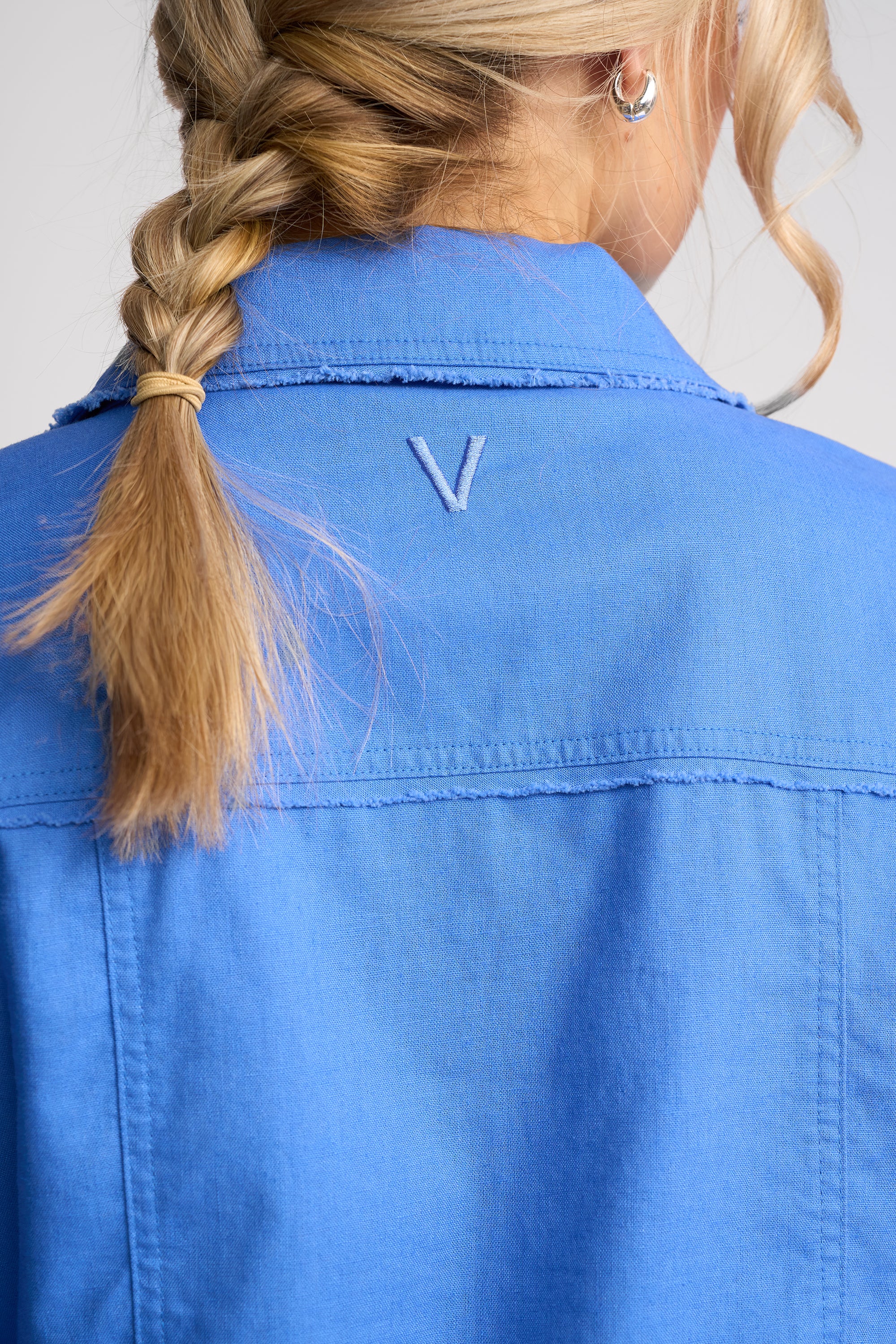 Close-up back view of a person wearing a bright blue collared shirt or jacket, with a small embroidered V-shaped logo centered below the collar. The person has blonde hair in a braid and is shown against a light neutral background.