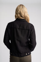 A woman viewed from behind wearing a black jacket and olive green pants against a light gray studio background.