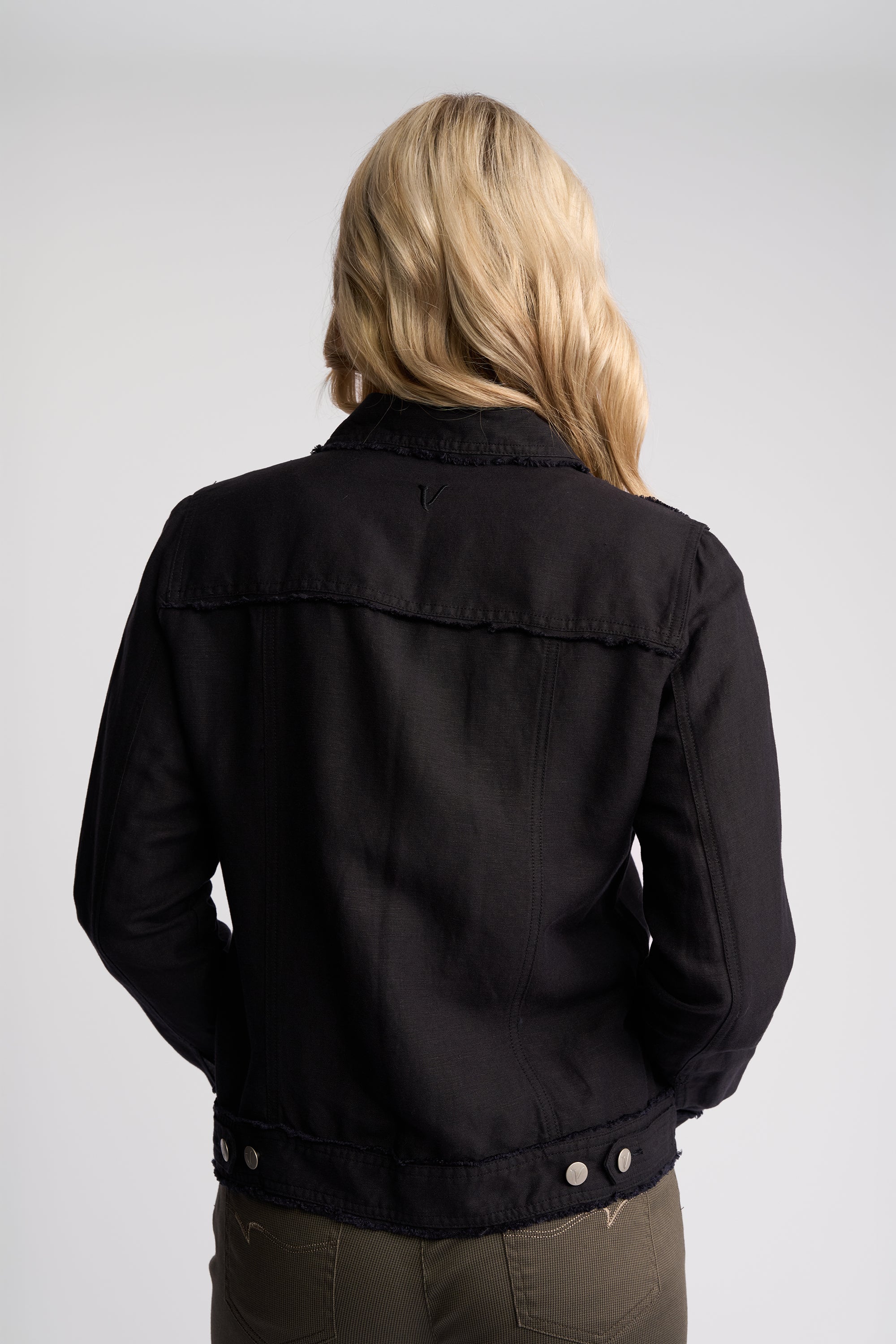 A woman viewed from behind wearing a black jacket and olive green pants against a light gray studio background.