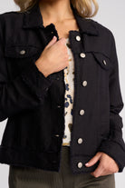 A close-up of a person wearing a black frayed denim jacket over a cream patterned top, with hands visible and a neutral studio background.
