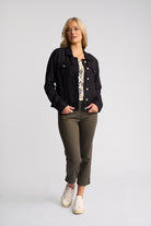 A woman modeling a black denim jacket over a patterned top, paired with olive green cropped pants and white sneakers, against a light gray studio background.