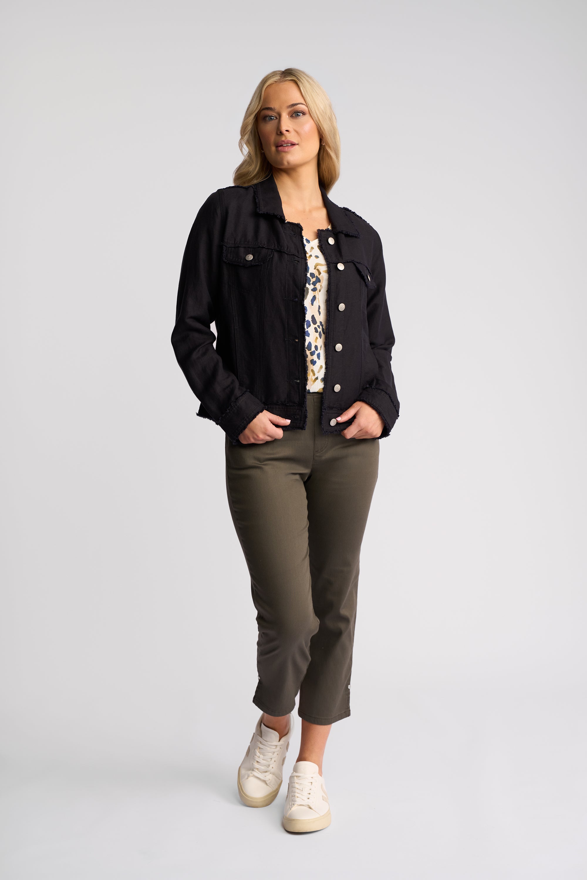 A woman modeling a black denim jacket over a patterned top, paired with olive green cropped pants and white sneakers, against a light gray studio background.