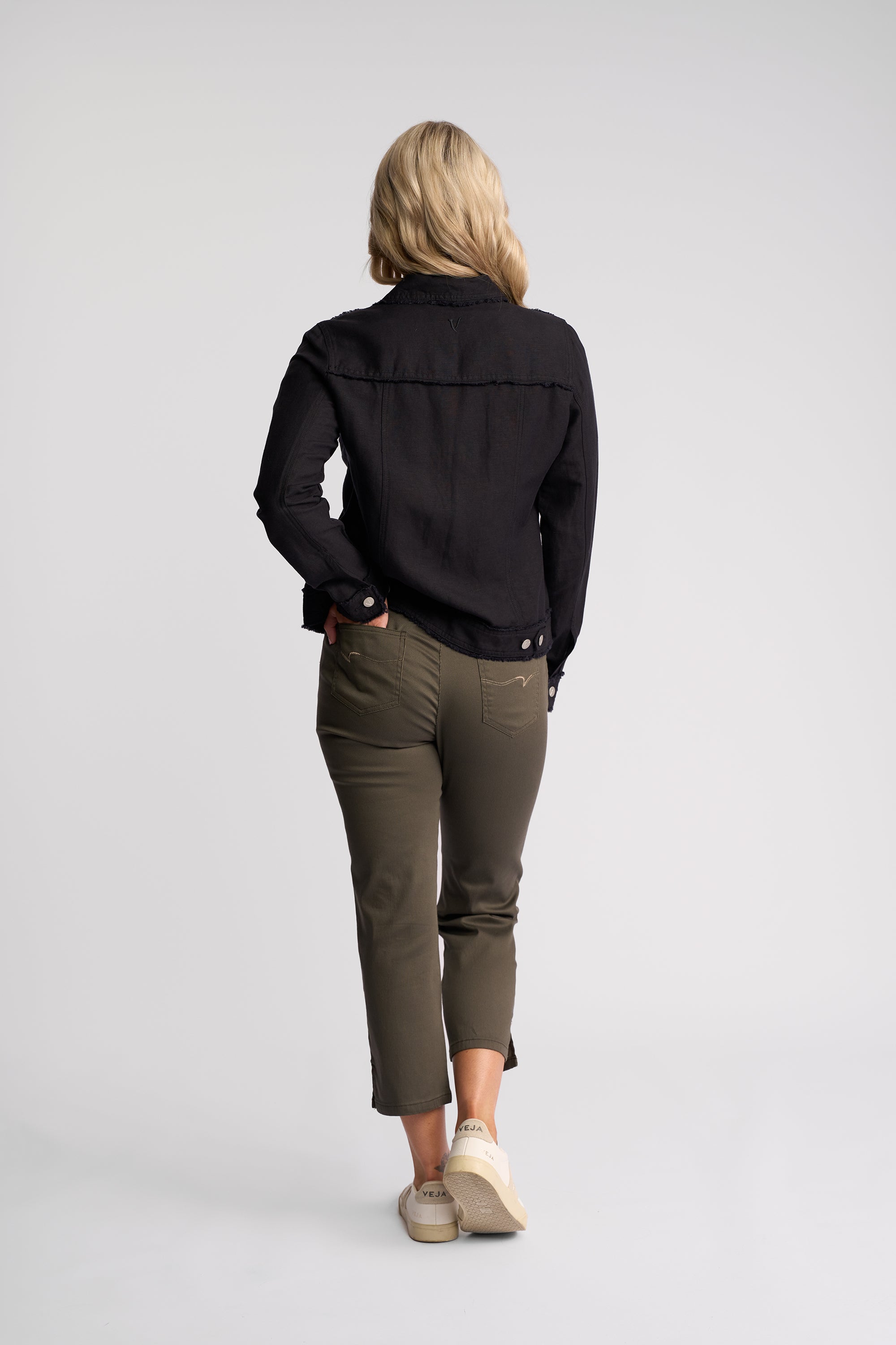 A woman is shown from behind wearing a black long-sleeve button-up shirt, olive green cropped trousers, and beige sneakers against a light gray studio background.