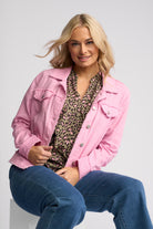 A woman wearing a light pink denim jacket over a leopard-print blouse and blue jeans, seated against a light gray studio background.