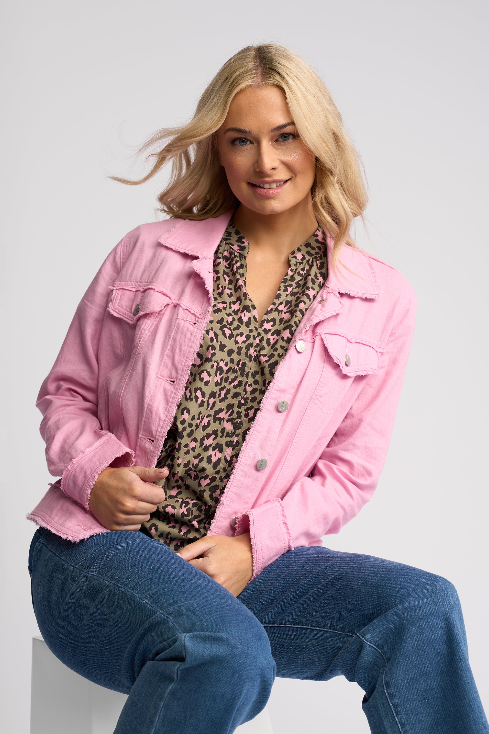 A woman wearing a light pink denim jacket over a leopard-print blouse and blue jeans, seated against a light gray studio background.