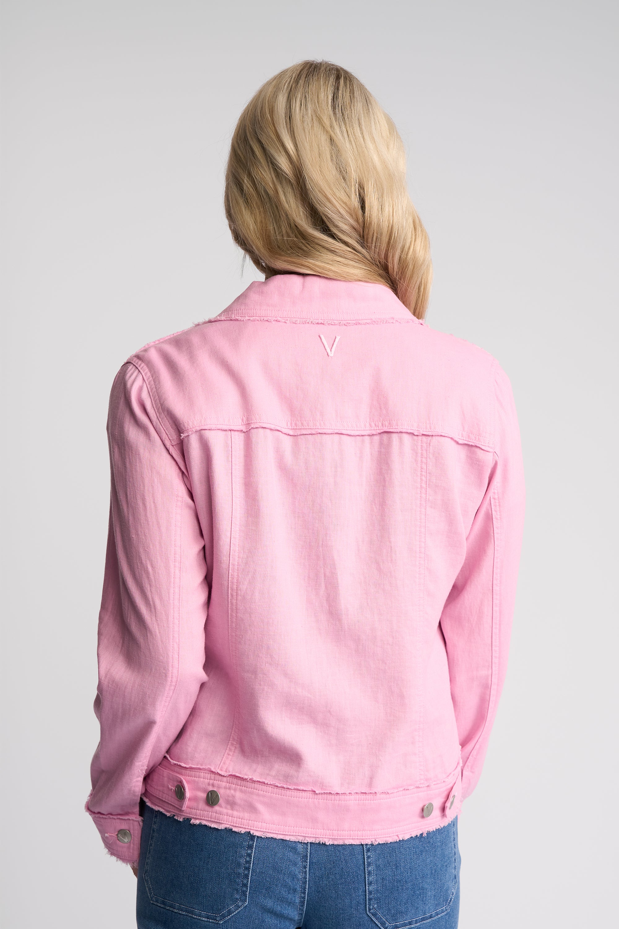 A woman viewed from behind wearing a light pink cropped denim jacket and blue jeans against a light gray studio background.