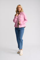A woman modeling a light pink denim jacket over a patterned top, paired with blue jeans and white sneakers, against a plain light gray studio background.