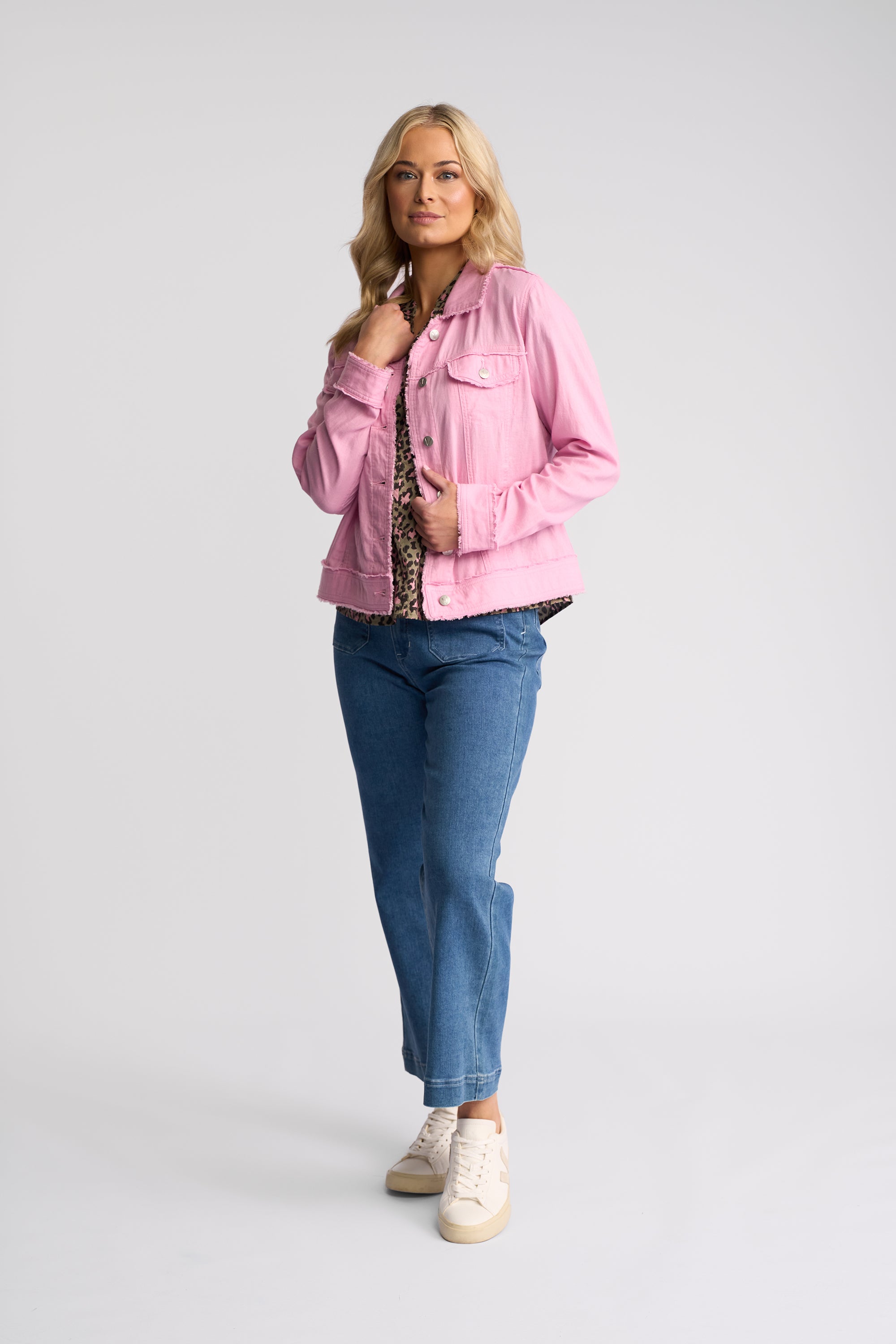 A woman modeling a light pink denim jacket over a patterned top, paired with blue jeans and white sneakers, against a plain light gray studio background.
