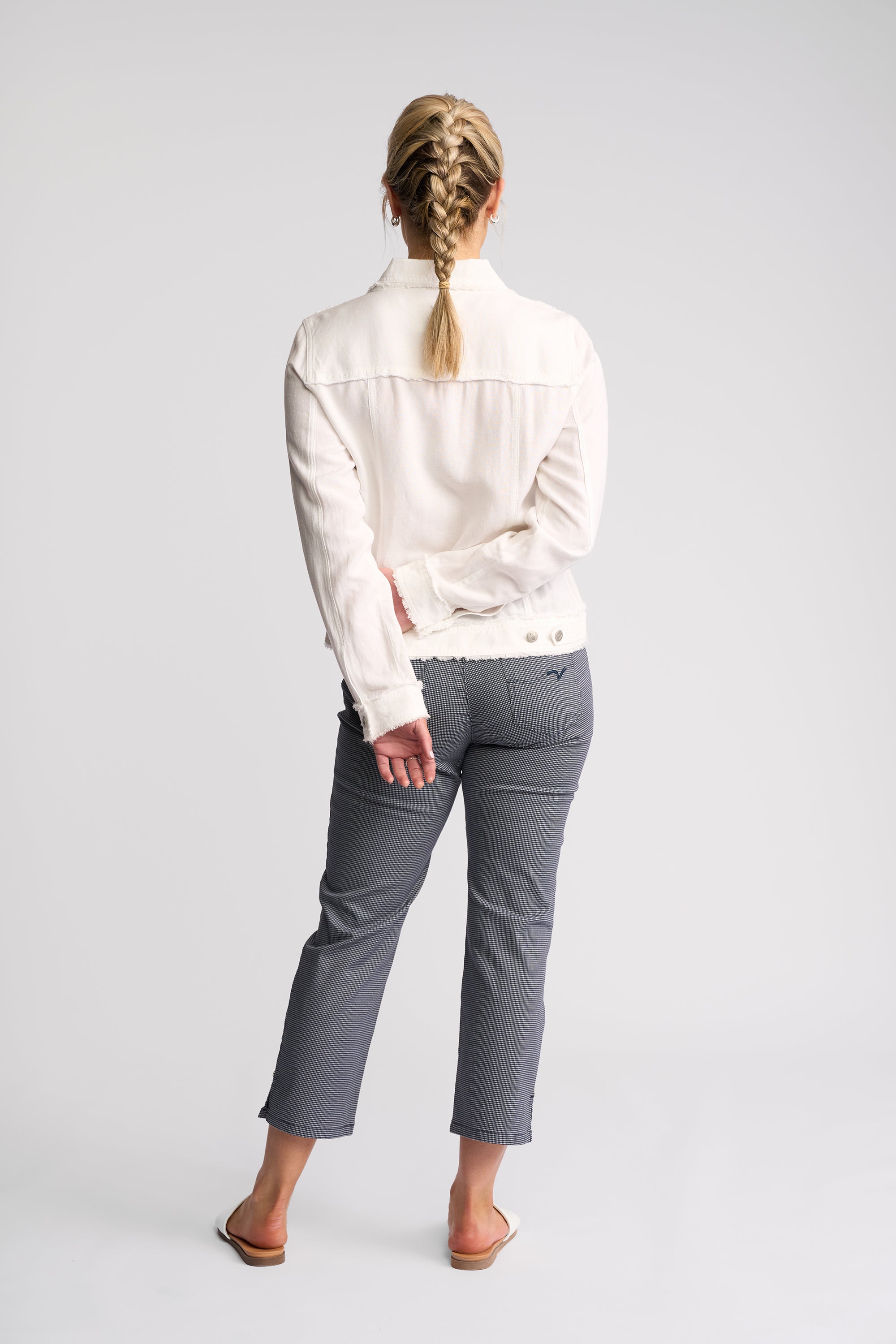 A woman shown from the back wearing a white long-sleeve jacket and gray cropped pants, standing against a light gray studio background.