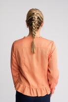 A woman viewed from behind wearing a peach-colored long-sleeve blouse with a ruffled neckline and hem, paired with dark navy pants, against a light gray studio background.