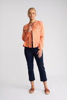A woman modeling a light peach-orange cropped denim jacket over a black patterned top, paired with dark navy cropped pants and brown slide sandals, against a light gray studio background.
