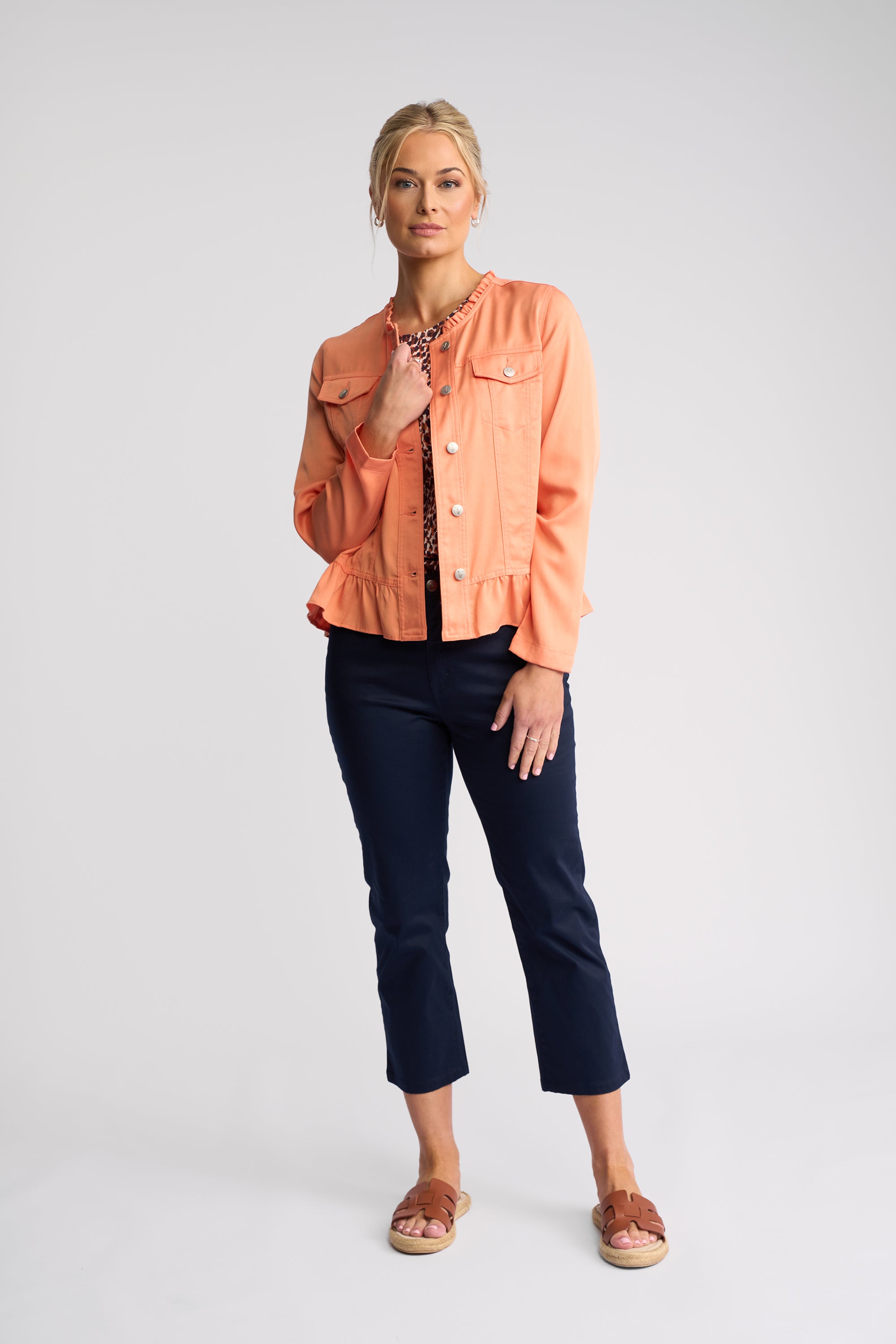 A woman modeling a light peach-orange cropped denim jacket over a black patterned top, paired with dark navy cropped pants and brown slide sandals, against a light gray studio background.