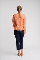 A woman viewed from behind wearing a peach-colored long-sleeve jacket with a ruffled hem and dark navy cropped pants, standing against a light gray studio background.