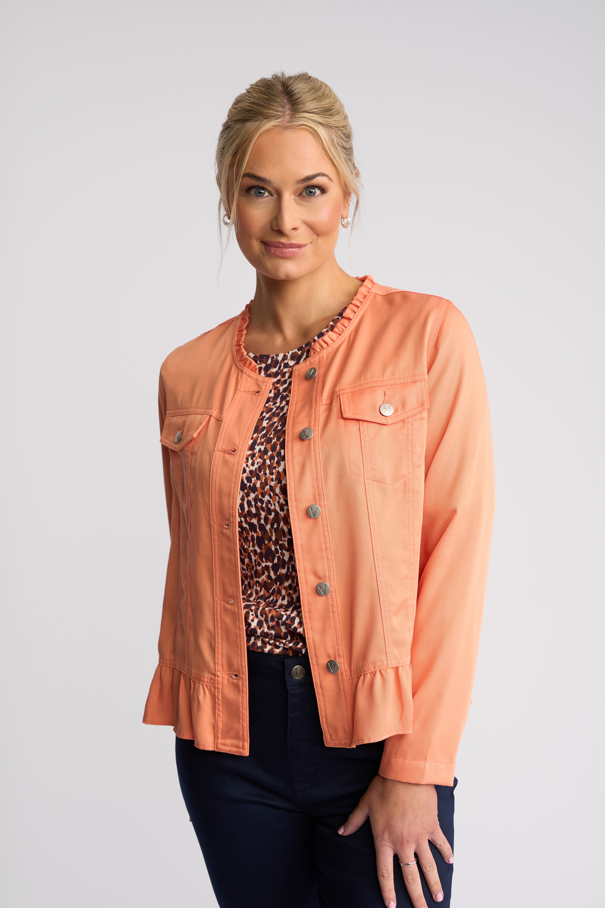 A woman modeling a coral peach cropped jacket with ruffle trim, button-front closure, and flap chest pockets, worn over a leopard-print top and dark pants against a light gray studio background.