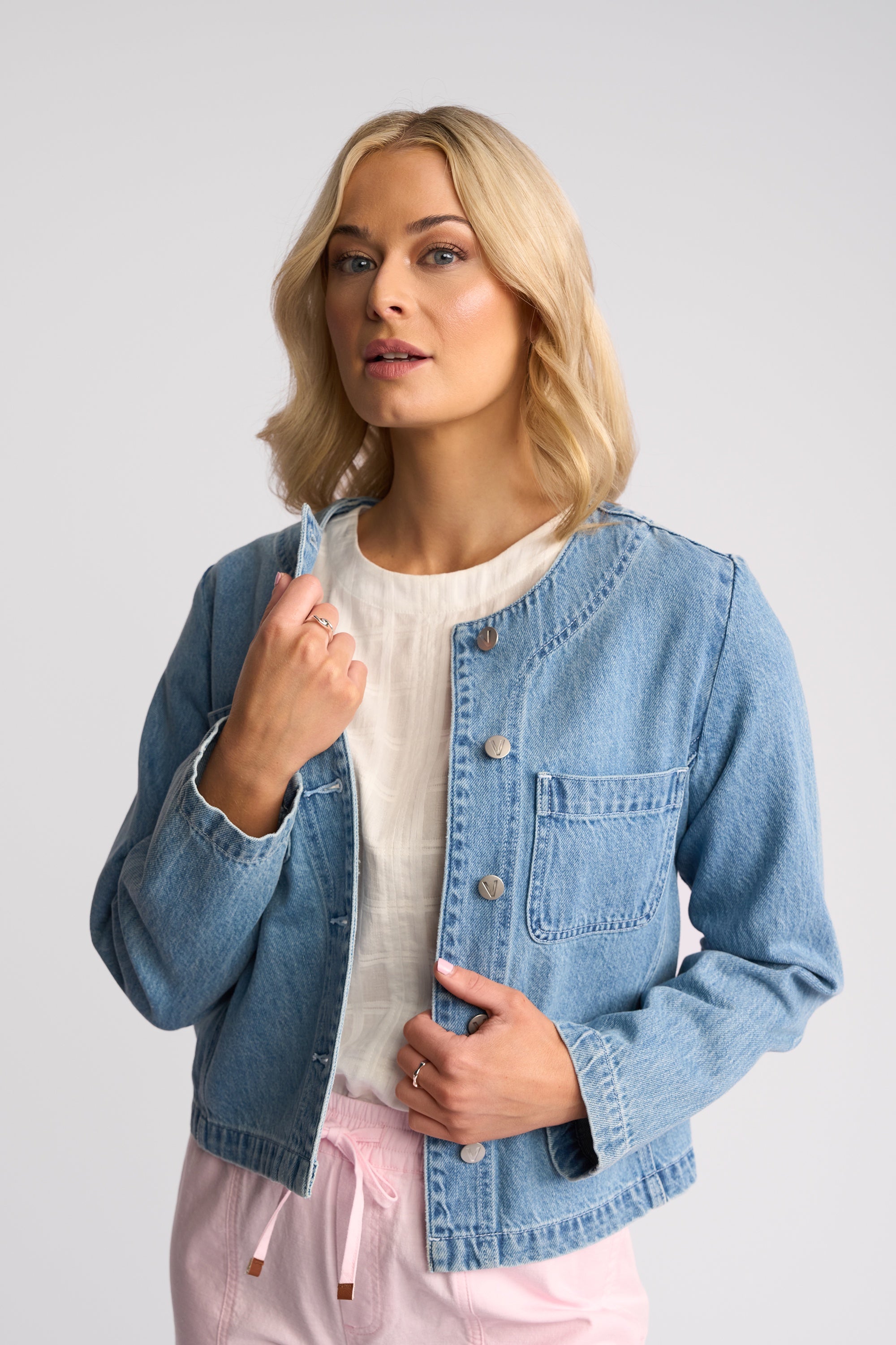 A blonde woman wearing a light blue cropped denim jacket over a white top and pink pants, posing against a light gray studio background.