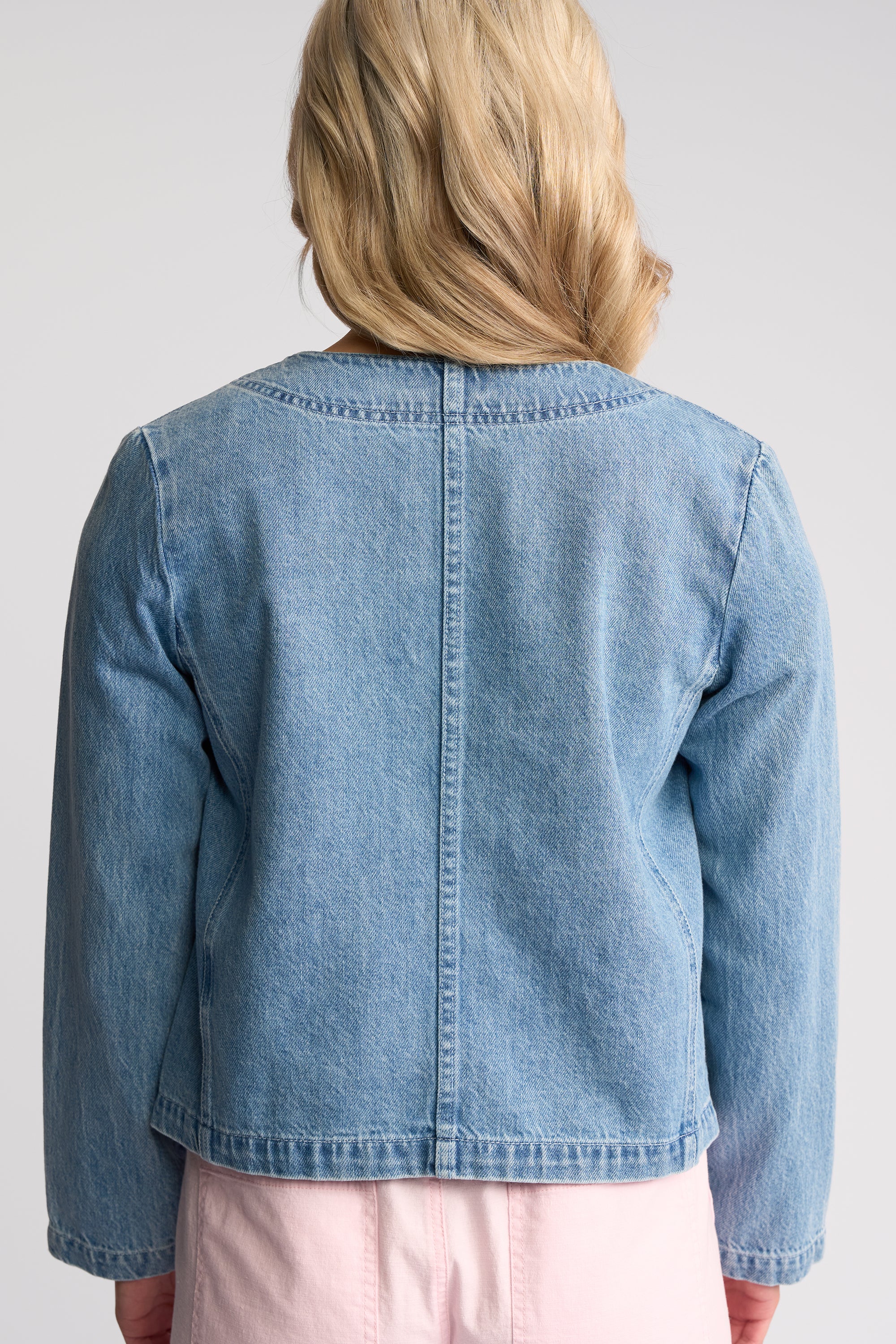 A child wearing a cropped light blue denim jacket, shown from the back against a light gray studio background.