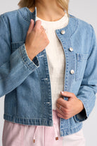 A woman wearing a cropped light blue denim jacket over a white top and pink drawstring pants, shown from the shoulders to the hips against a light gray studio background.